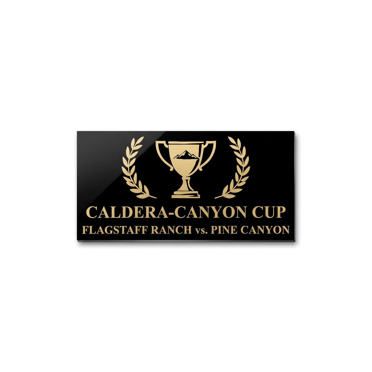 A rectangular black plaque or sign with gold lettering reading 'CALDERA-CANYON CUP' and 'FLAGSTAFF RANCH vs. PINE CANYON,' centered around a gold trophy icon with laurel branches, shown on a plain white background.