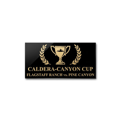 A rectangular black plaque or sign with gold lettering reading 'CALDERA-CANYON CUP' and 'FLAGSTAFF RANCH vs. PINE CANYON,' centered around a gold trophy icon with laurel branches, shown on a plain white background.