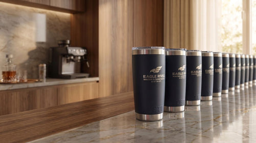 A row of dark navy blue insulated travel tumbler cups with silver metal rims and lids, lined up on a polished marble countertop in a modern kitchen setting. The row of cups feature the text 'EAGLE RIVER WATER & SANITATION DISTRICT' along with their eagle organization logo.
