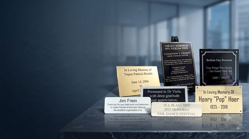 A group of personalized memorial plaques and engraved metal plates displayed on a dark reflective surface against a blurred dark blue background. The engraved metal plaques include gold, silver, and black options with engraved text such as 'In Loving Memory of ' , 'Thank you' and 'Presented to Dr.'