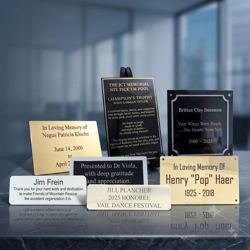 A group of personalized memorial plaques and engraved metal plates displayed on a dark reflective surface against a blurred dark blue background. The engraved metal plaques include gold, silver, and black options with engraved text such as 'In Loving Memory of ' , 'Thank you' and 'Presented to Dr.'