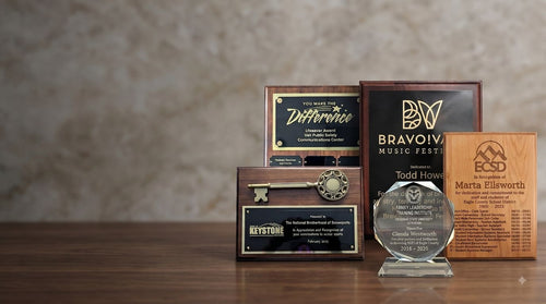 A collection of engraved wooden and acrylic awards and plaques displayed on a wooden table against a textured beige wall. The set includes a round clear glass award on a base, a black and gold 'Bravo! Vail Music Fest' plaque, a brown wood plaque, a smaller brown wood plaque, and a dark wood plaque with a key detail.