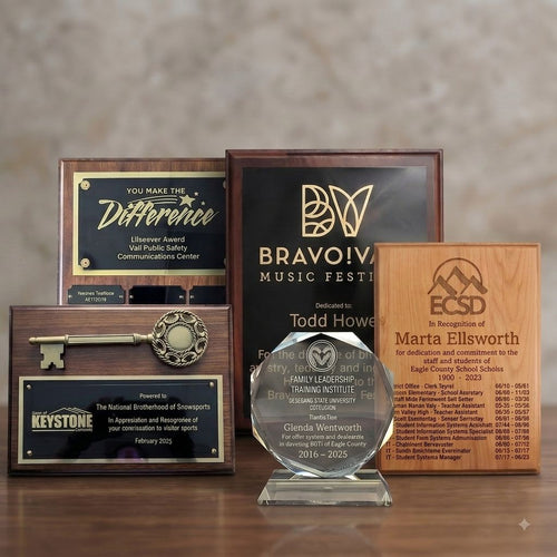 A collection of personalized award plaques and trophies displayed on a wooden surface against a neutral background. The set includes a large black and gold Bravo! Music Festival trophy, a wooden plaque for Eagle County School District, a wooden Keystone Colorado award, a glass crystal family leadership trophy, and a decorative key on a wooden base.