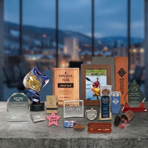 A collection of custom corporate awards and trophies displayed on a gray concrete table in front of a blurred office window background. The trophies include a blue and gold trophy cup, a wooden 'Employee of the Year' plaque, a photo frame, a clear acrylic award, a pink star, and several small metal and wooden keepsakes.