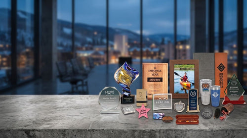 A collection of custom corporate awards and trophies displayed on a gray concrete table in front of a blurred office window background. The trophies include a blue and gold trophy cup, a wooden 'Employee of the Year' plaque, a photo frame, a clear acrylic award, a pink star, and several small metal and wooden keepsakes.